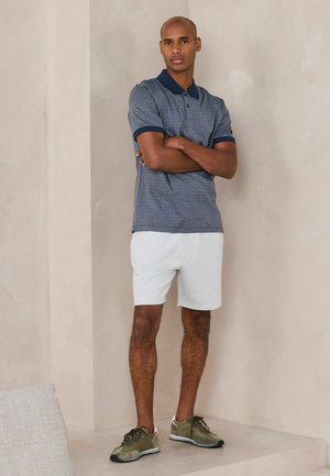 Muž stojaci so skríženými rukami, oblečený v modré vzorované polo, biele šortky a zelené tenisky pred béžovou stenou.