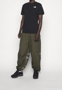 Camiseta negra de manga corta con logo blanco, pantalones cargo verdes oliva con diseños gráficos blancos y zapatillas negras en una persona de pie.