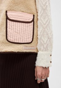 Ταν γιλέκο σε σέρπα ύφασμα με ροζ καπιτονέ τσέπη, καφέ φινίρισμα και ετικέτα με το λογότυπο. Λευκός υφασμάτινος μανίκας με κουμπιά ως διακοσμητικά και καφέ ριμπ φούστα.