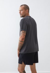 Athletisches Shirt aus dunkelgrauem Stoff mit Netz-Paneelen. Kombiniert mit schwarzen Shorts mit seitlichen Schlitzen und Logo-Details.