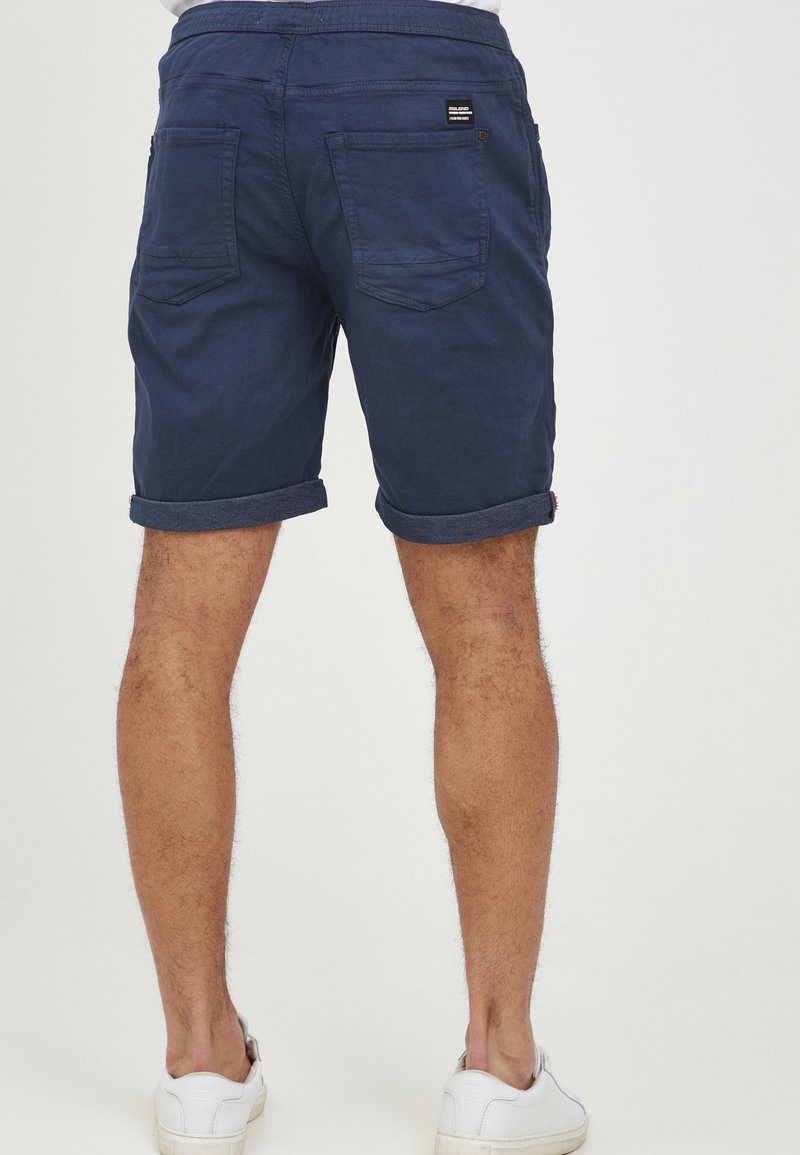 Shorts en coton bleu marine avec revers roulés, deux poches arrière, et une petite étiquette de marque. Texture lisse et coupe ajustée.