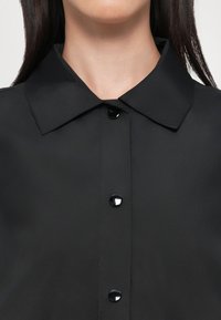 Camicia nera con bottoni a pressione, caratterizzata da un colletto classico, bottoni neri lucidi e una trama della stoffa liscia e opaca. Design semplice e lineare.
