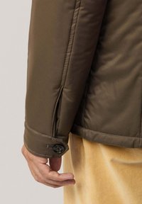 Chaqueta acolchada marrón con una textura suave, que cuenta con un cierre de botón en el puño. El tejido parece ser resistente al agua y ligero.