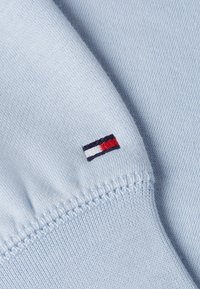 Tessuto di cotone azzurro chiaro con un piccolo logo ricamato che presenta strisce rosse, bianche e blu vicino alla cucitura. Texture morbida, cuciture visibili.