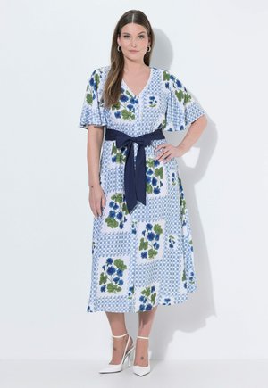 Vrouw draagt een midi-jurk met blauw-wit bloemmotief, een marineblauwe tailleband en witte hoge hakken, staand tegen een witte achtergrond.