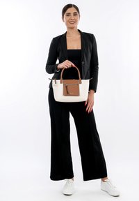 Sac en toile avec un corps crème et des accents en cuir marron, équipé d'une poche zippée à l'avant et de double poignées. Modèle vêtu d'une tenue noire.