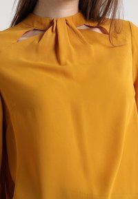 Senapsgul blus med en mjuk, slät textur, som har hög krage, utskärningar och en rosettknut vid halsringningen.