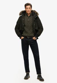 Veste noire rembourrée avec capuche en fausse fourrure, pull en maille verte, jeans foncés et bottes grises. Comprend une montre et une ceinture noire.