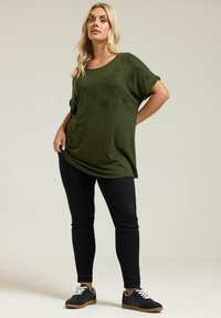 Camiseta oversized verde oliva con estampado de estrellas negras, combinada con jeans skinny negros y zapatillas negras con cordones blancos y suelas marrones.