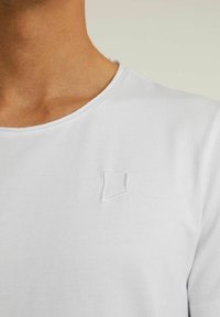Tricou alb din bumbac, cu guler rotund și mâneci scurte, având un mic logo geometric brodat pe piept.