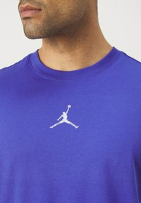 Camiseta de algodón morada con escote redondo, que presenta un logo de Jumpman blanco en el pecho. Textura suave y mangas cortas.