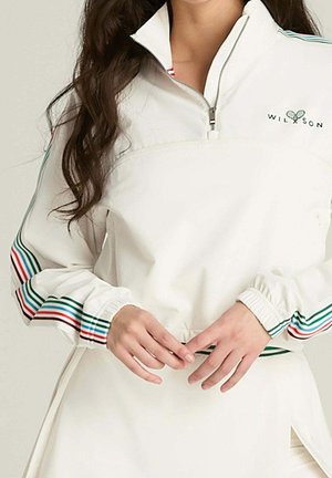 Frau trägt einen weißen langärmligen Pullover mit Reißverschluss und bunten gestreiften Details an Ärmeln und Bund, Hände vor der Taille verschränkt.