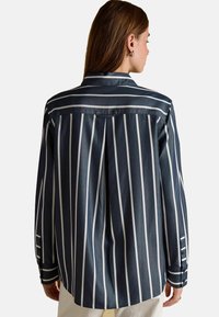 Naine, kes kannab tumesinist ja valgeid vertikaalselt triibulisi pikkade varrukatega särki, vaadates seljaga, kergelt heledates pükstes.