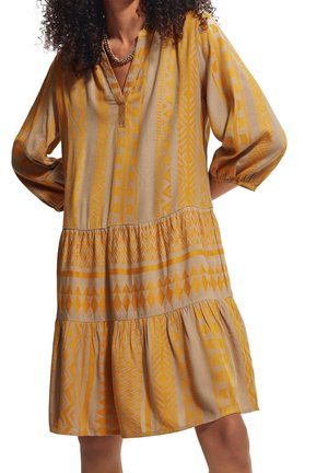 Mujer con vestido beige suelto hasta la rodilla con patrones geométricos naranjas y mangas abullonadas tres cuartos, con las manos apoyadas en las caderas.