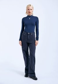 Dunkelblaues Langarmoberteil kombiniert mit hoch taillierten, locker sitzenden schwarzen Jeans; Modell trägt schwarze Schuhe; minimale sichtbare Accessoires.