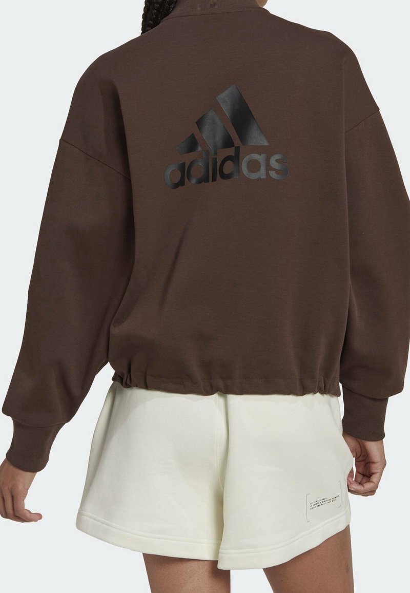 Sudadera de forro polar marrón con dobladillo elástico y diseño oversize, que presenta un logo de Adidas negro y brillante en la parte trasera, combinada con pantalones cortos en color blanco roto.