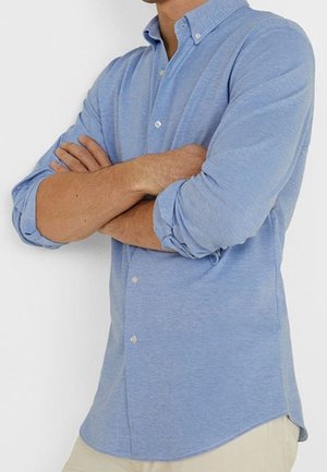 Mężczyzna stojący z założonymi ramionami, ubrany w jasnoniebieską koszulę na guziki z podwiniętymi rękawami oraz beżowe spodnie na jednolitym tle.