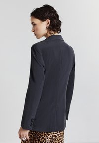 Femme aux cheveux courts et foncés portant un blazer ajusté noir et une jupe à imprimé léopard, tournée sur le côté contre un fond clair et uni.