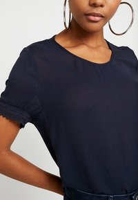 Navy blue short-sleeve top with a round neckline and lace trim on sleeves, featuring a lightweight fabric and subtle texture.