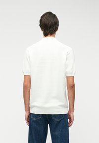 Chemise polo blanche à manches courtes avec une surface texturée et un col côtelé, portée avec un jean en denim bleu, vue de dos.