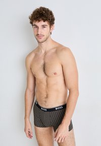 Schwarze Boxer Shorts mit weißem Punktemuster und 'BOSS'-Logo am Bund. Das Model hat kurze lockige Haare, ist oberkörperfrei und posiert seitlich.