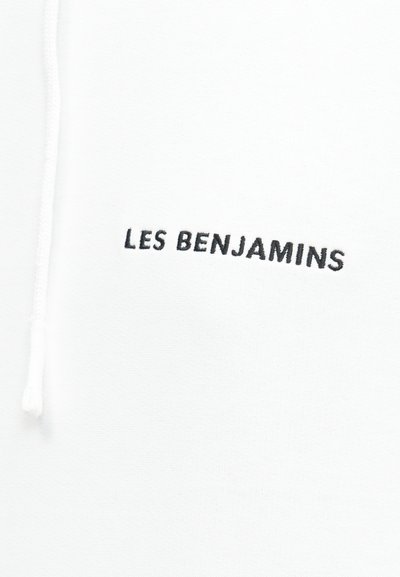 Primer plano de tela blanca con texto bordado en negro "LES BENJAMINS" y un cordón blanco de ajuste en el lado izquierdo.