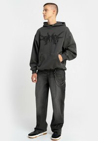 Czarna, oversize bluza z kapturem z graficznym nadrukiem, kieszenią z przodu, w połączeniu z ciemnymi dżinsami z detalem w postaci łańcucha oraz czarnymi trampkami z białą podeszwą.