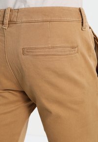 Béžové bavlněné chino kalhoty s jednou zadní kapsou s obdélníkovým klopou, hladkým povrchem a prošitím podél pasu a okraje kapsy.