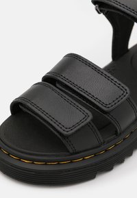 Black sandals made of textured synthetic material, featuring three adjustable straps and a thick, ridged sole with yellow stitching accents.