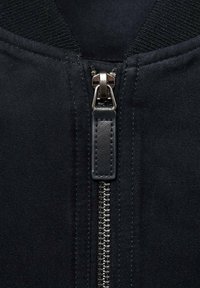 Chaqueta negra con cremallera que presenta un brillo plateado, tela texturizada alrededor del cuello y una superficie suave con detalles de costura mínimos.