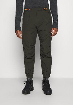 Mężczyzna ubrany w ciemnozielone spodnie dresowe z elastycznymi mankietami, czarne buty trekkingowe i ciemną bluzę z długim rękawem na jednolitym tle.