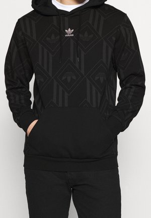 Osoba nosząca czarną bluzę z kapturem Adidas z subtelnym geometrycznym wzorem i przednią kieszenią, ręce częściowo w kieszeni.
