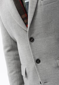 Blazer grigio in lana con rever strutturati, due bottoni neri e una tasca sul petto che rivela un rivestimento a motivo in tonalità di rosso e verde.