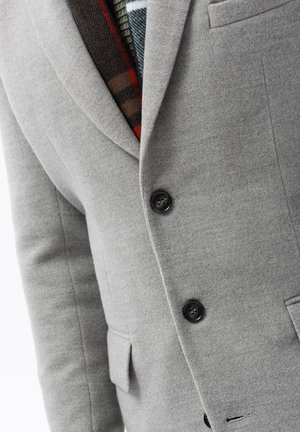 Grau wollene Blazer mit strukturierten Revers, zwei schwarzen Knöpfen und einer Brusttasche, die ein gemustertes Futter in Rot- und Grüntönen zeigt.