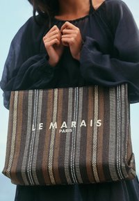 Textured tote bag in brown, cream, and black stripes. Features logo "LE MARAIS PARIS" in white, with subtle patterns and a spacious design.