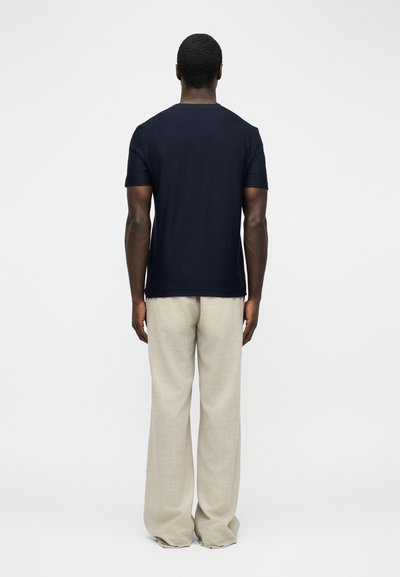 Άνδρας που στέκεται με την πλάτη προς την κάμερα, φορώντας μια ναυτική κοντομάνικη μπλούζα και χαλαρό μπεζ παντελόνι, μπροστά από απλό φόντο.