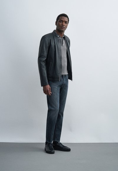 Νεαρός άνδρας στέκεται μπροστά σε απλό φόντο, φοράει μαύρο δερμάτινο μπουφάν, γκρι πουκάμισο, σκούρα τζιν και μαύρα αθλητικά παπούτσια.