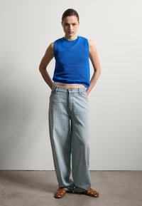 Hellblau gestricktes ärmelloses Oberteil, hellblaue weit geschnittene Jeans und braune geflochtene Sandalen. Glatte Textur und lockere Passform. Schlichtes Design ohne Muster.