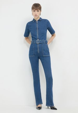 Frau in einem enganliegenden Jeans-Overall mit einem Reißverschluss vorne, Gürtel, kurzen Ärmeln und schwarzen spitzen Absatzschuhen vor einem schlichten weißen Hintergrund.