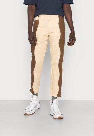 Man die beige en bruine colorblock-broek draagt, grijze sokken, witte hoge sneakers en een marineblauw shirt, staand tegen een effen muur.