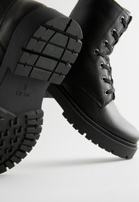 Botas negras con cordones, hechas de cuero texturizado, que cuentan con una suela de goma resistente con profundos surcos para mayor tracción. Ojetes reforzados y cordones redondos.