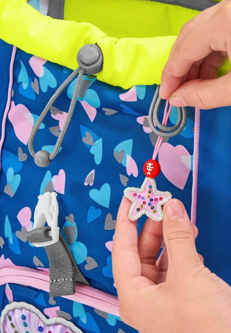 Children's backpack with a blue fabric featuring pink and gray heart patterns. A star-shaped zipper pull is held, adorned with colorful beads.