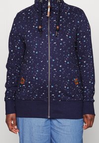 Sudadera con cremallera en color marino con un patrón floral en azul claro y blanco. Tiene un cuello alto, cordones ajustables y bolsillos laterales.