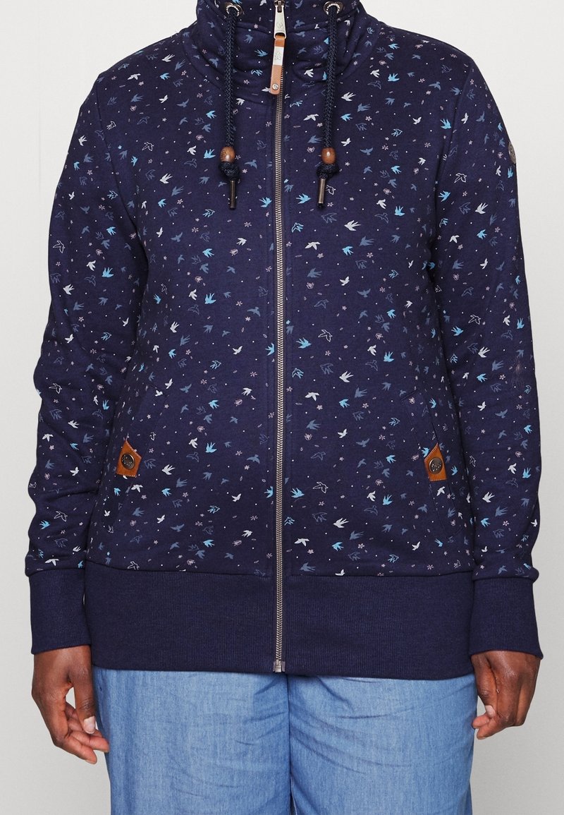 Sudadera con cremallera en color marino con un patrón floral en azul claro y blanco. Tiene un cuello alto, cordones ajustables y bolsillos laterales.