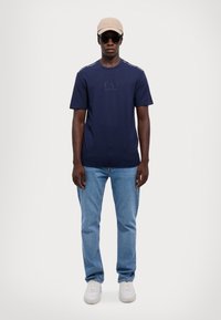 Marineblauw t-shirt met een ronde hals en logo, lichtblauwe jeans, witte sneakers, een baseballpet en zonnebril. Casual, relaxte pasvorm.