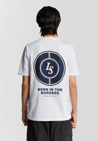 Hvid bomulds T-shirt med et stort cirkulært grafisk motiv i marineblå og guld på ryggen, der indeholder teksten "BORN IN THE BORDERS" samt brandingdetaljer.