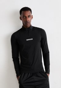 Mann iført svart langermet Calvin Klein genser med halv glidelås og svarte bukser, stående mot en enkel hvit vegg med hendene i lommene.