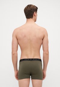Olivgrüne Boxershorts mit einem schwarzen Elastikbund, auf dem der Text "PIER ONE" in einem sich wiederholenden Muster zu sehen ist, von hinten gezeigt.