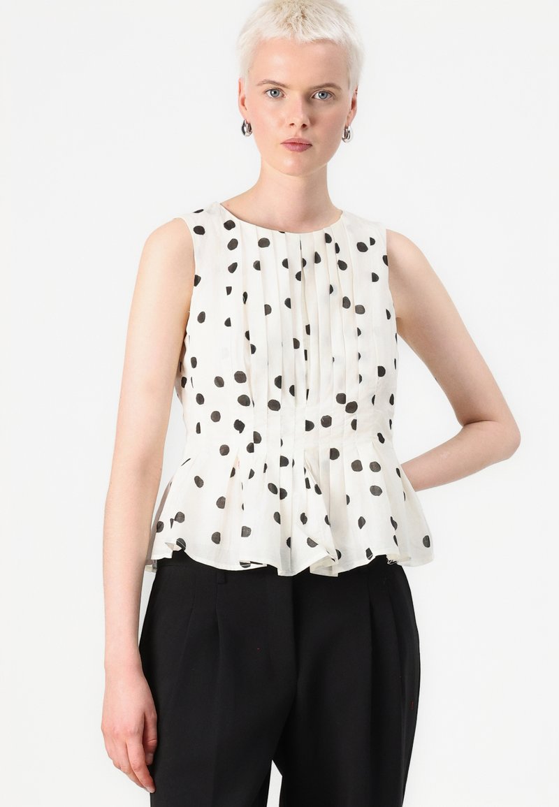 Haut sans manches plissé en crème avec des pois noirs. Présente une taille peplum et un col rond, conçu avec un tissu léger.
