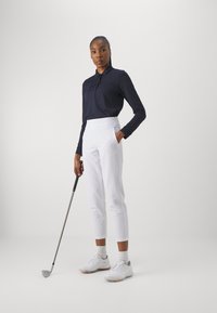 Polo shirt à manches longues marine, pantalons tailleur blancs taille haute et chaussures de golf blanches. Tenant un club de golf, fond neutre.